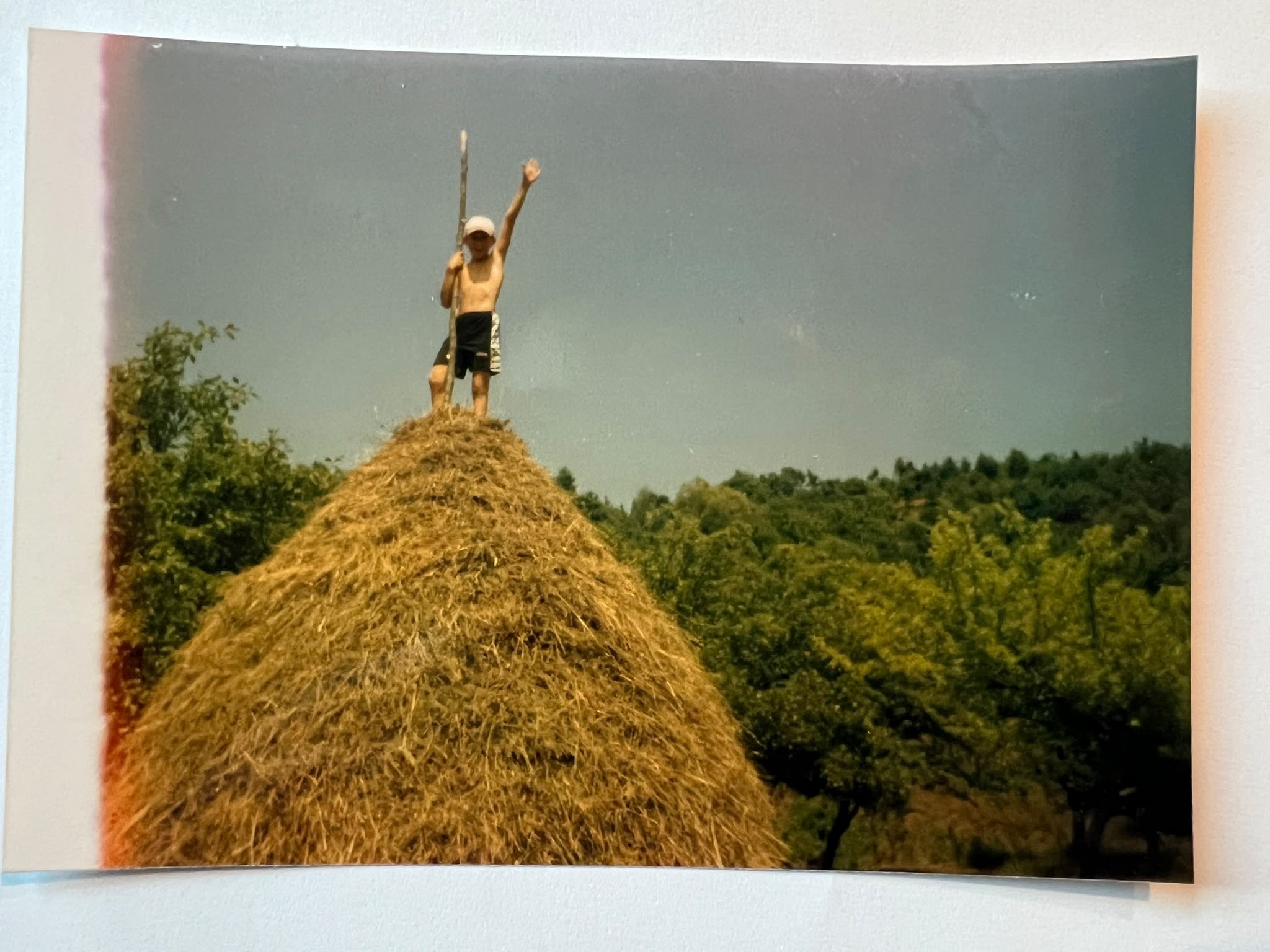 A boy stands on top of a tall haystack with his arms spread wide, against a hazy sky and green hills.