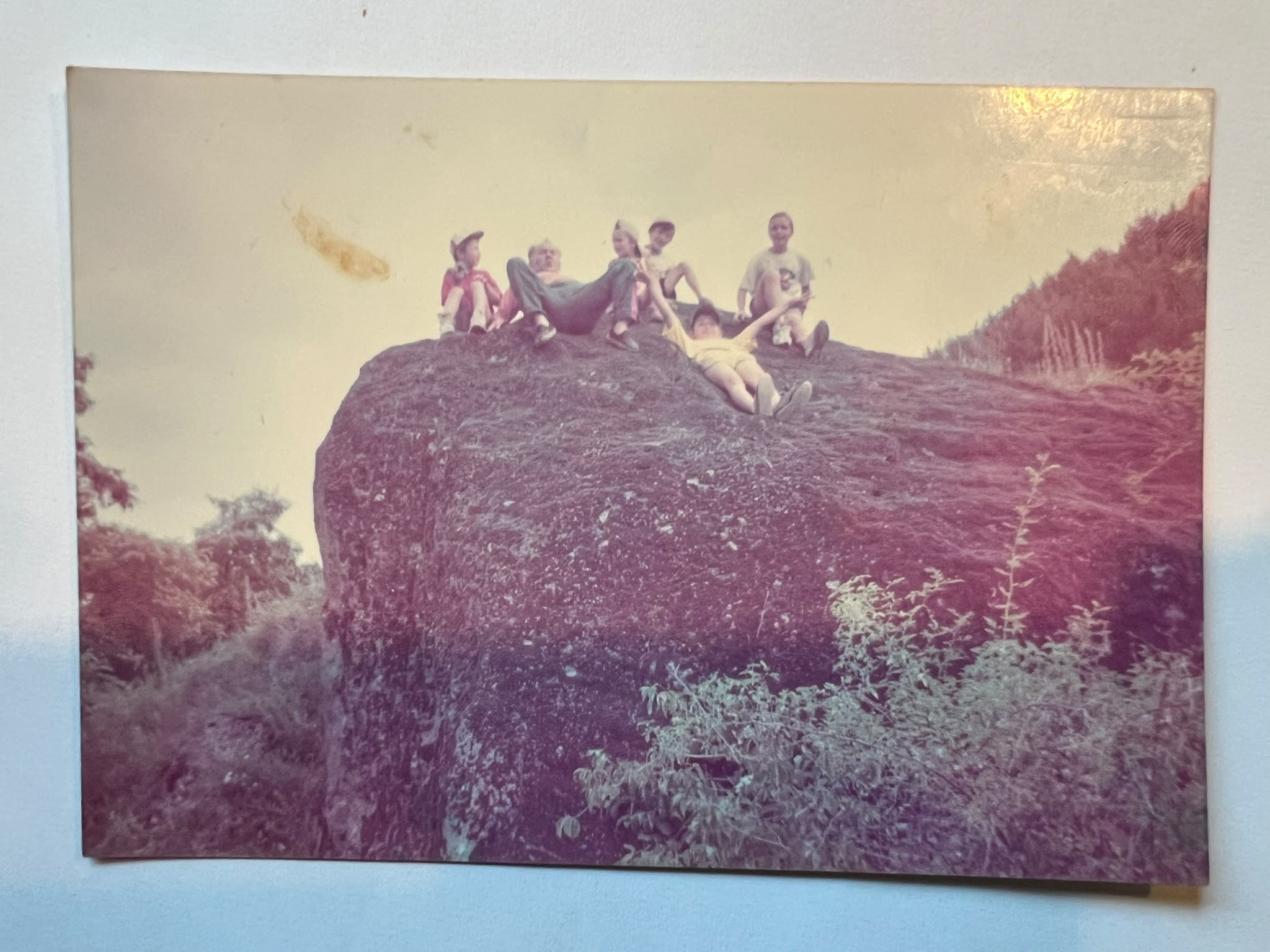 A group of children and a young man sit and lie on top of a large boulder against the sky.