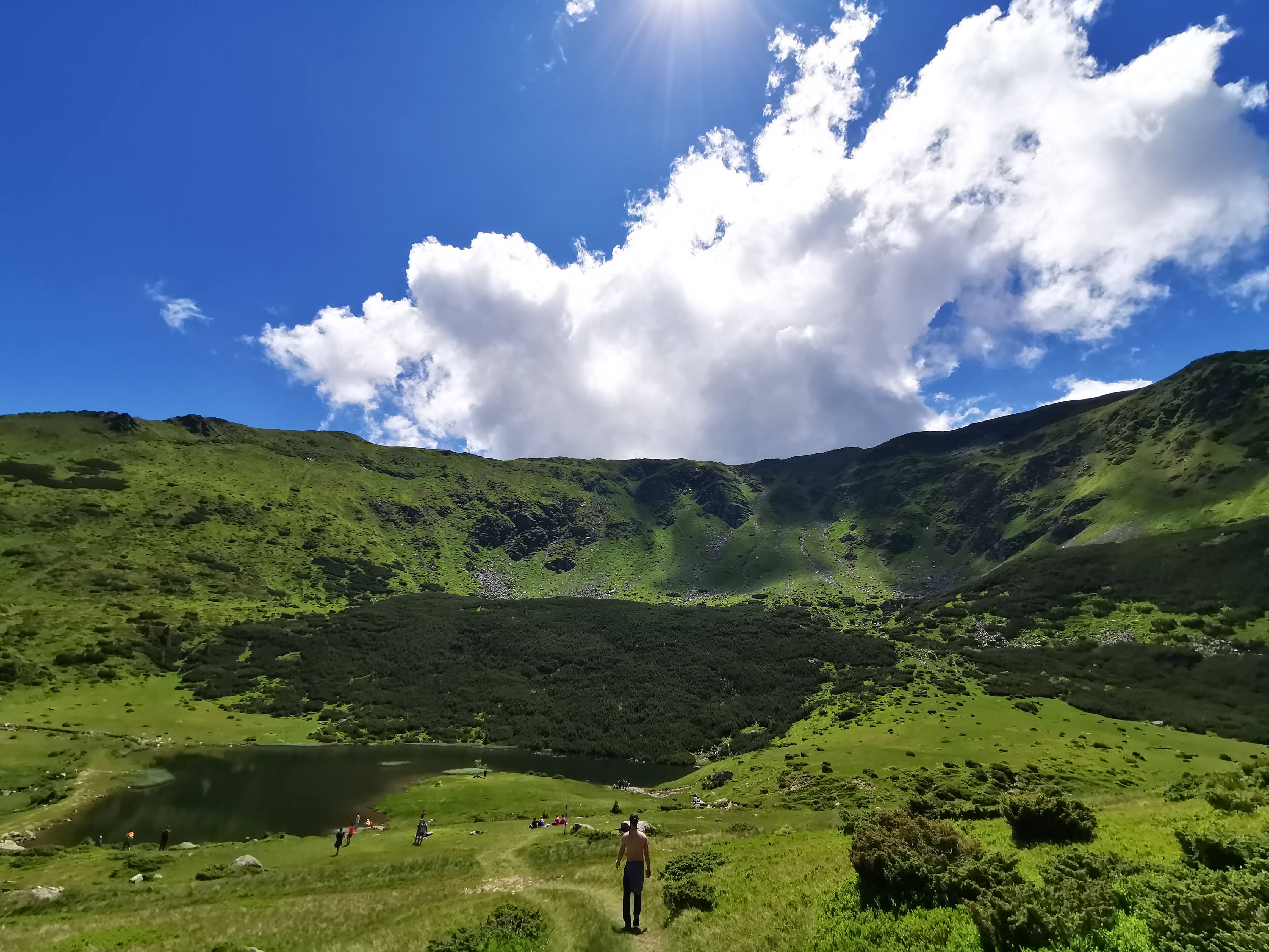 A lone walker descends toward a mountain lake beneath a bright sky and large clouds.
