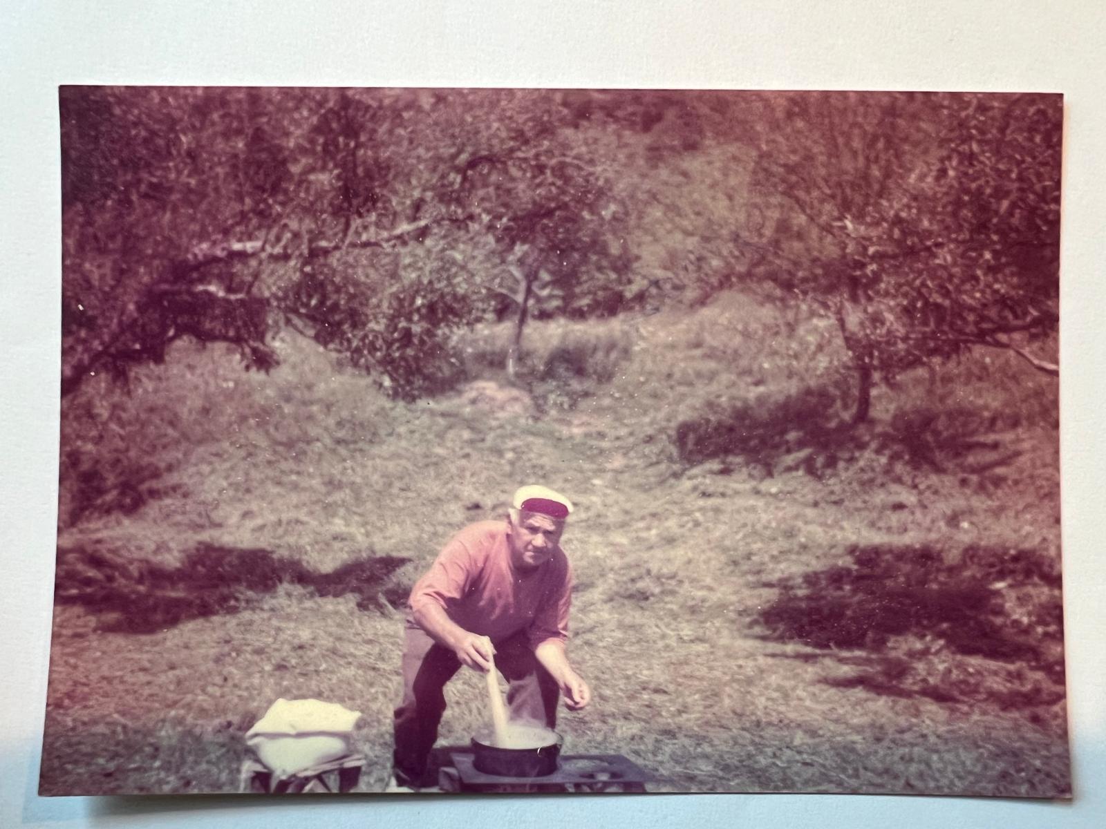 An older man in a white cap leans over a large pot on an outdoor stove, stirring it with a wooden spoon in an orchard.