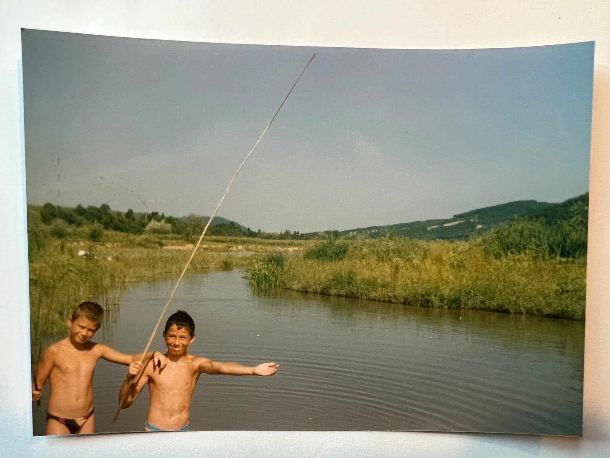 Two shirtless boys stand by a lake, one holding a long thin rod, with reeds and hills behind them.